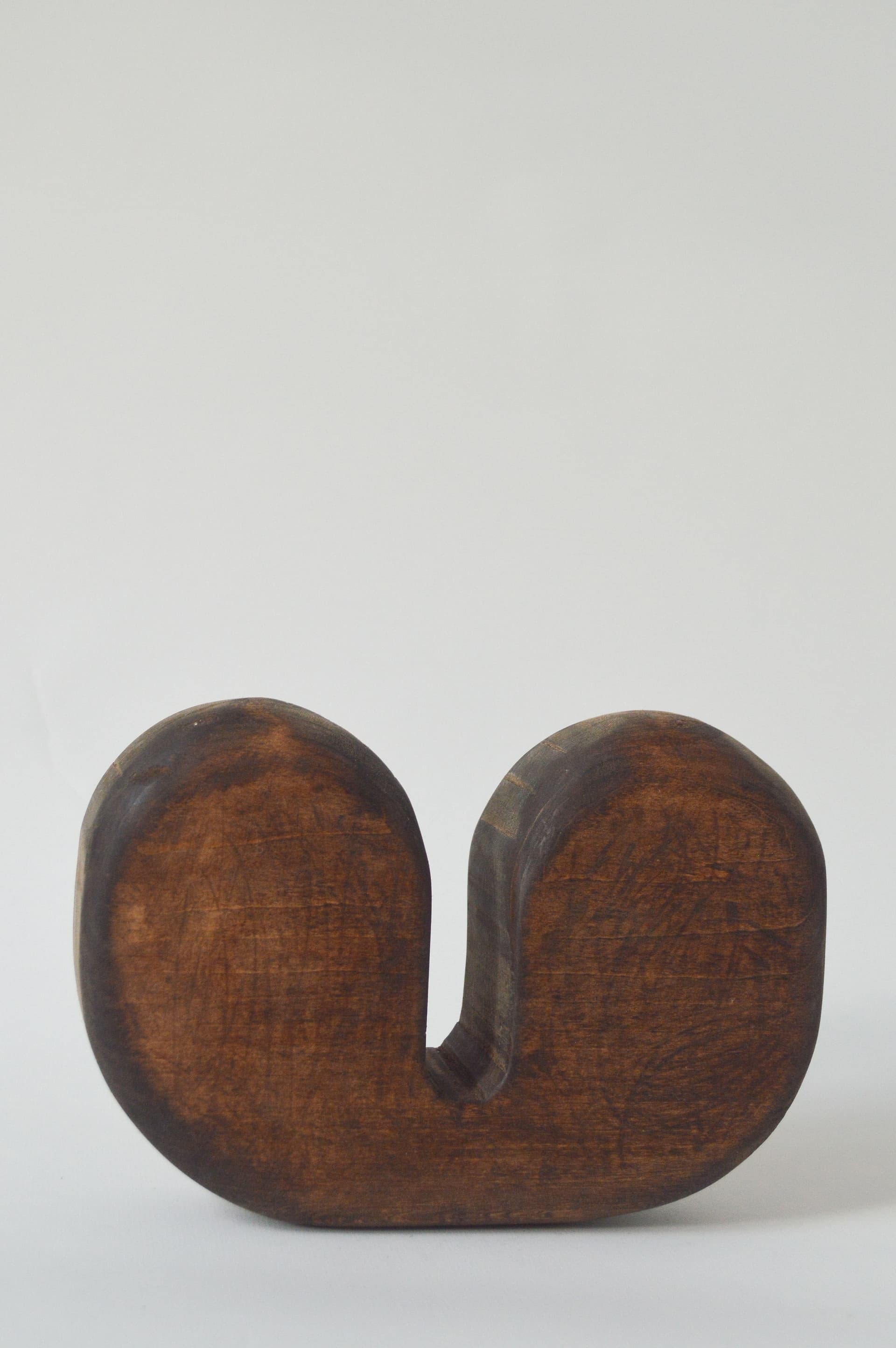 A wooden sculpture with a smooth, dark brown finish, shaped like a rounded "W" or a curvy "U" on its side, set against a plain white background.