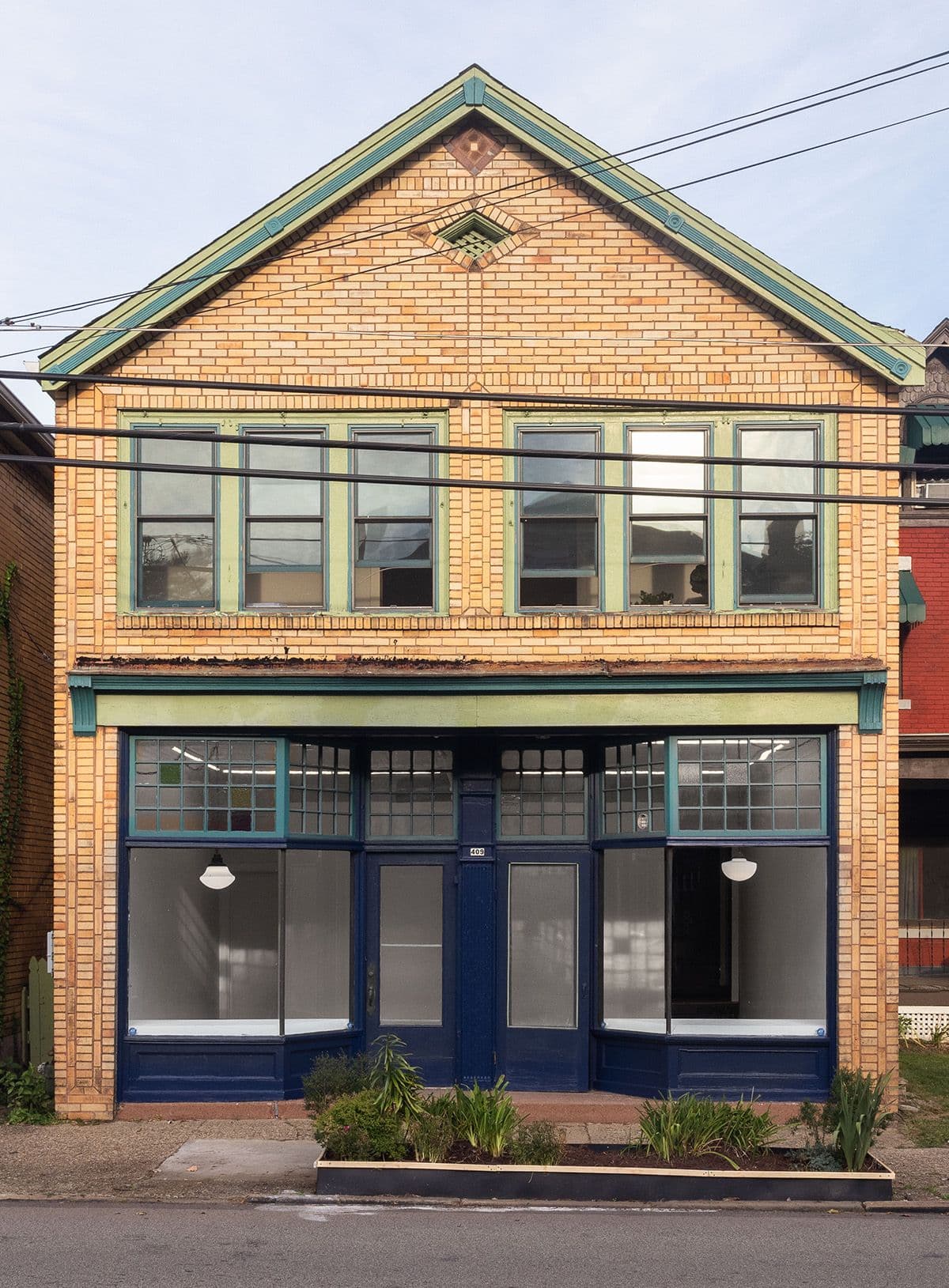 A two-story brick building with a slanted roof, featuring large glass windows on both floors. The lower level has double doors and shopfront windows with dark blue frames. The building is bordered by other structures and has plants along the sidewalk.