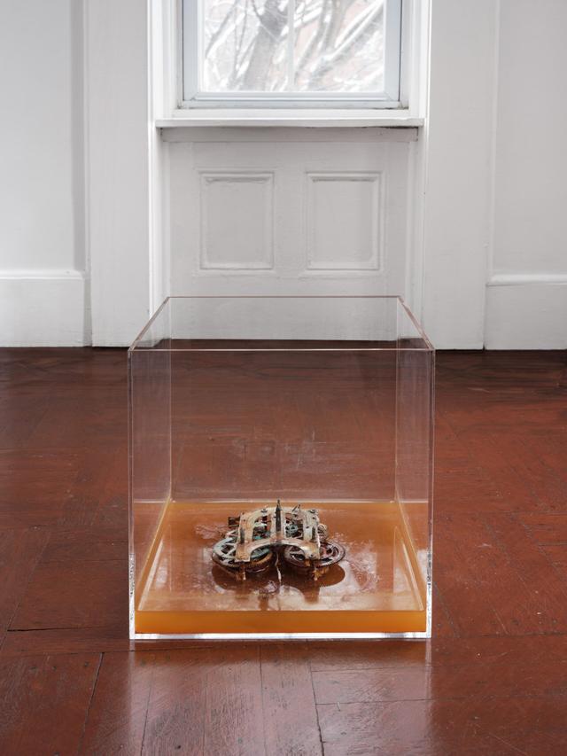 Micah Schippa. The last sentence ever written, 2024. Brass clock movement mechanism in gastric solution (HCL solution made to mimic the composition of stomach acid), plexiglass vitrine. 15 x 15x 15 inches.