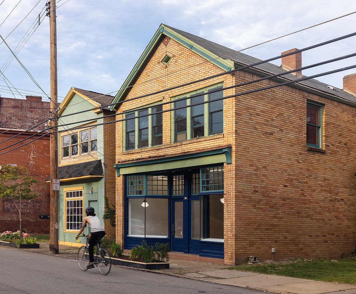 A cyclist rides past two adjacent two-story buildings on a quiet street. The buildings have distinctive features: one with a light green facade and the other with a brick exterior and blue trim around its large windows and entrance. Power lines cross overhead.
